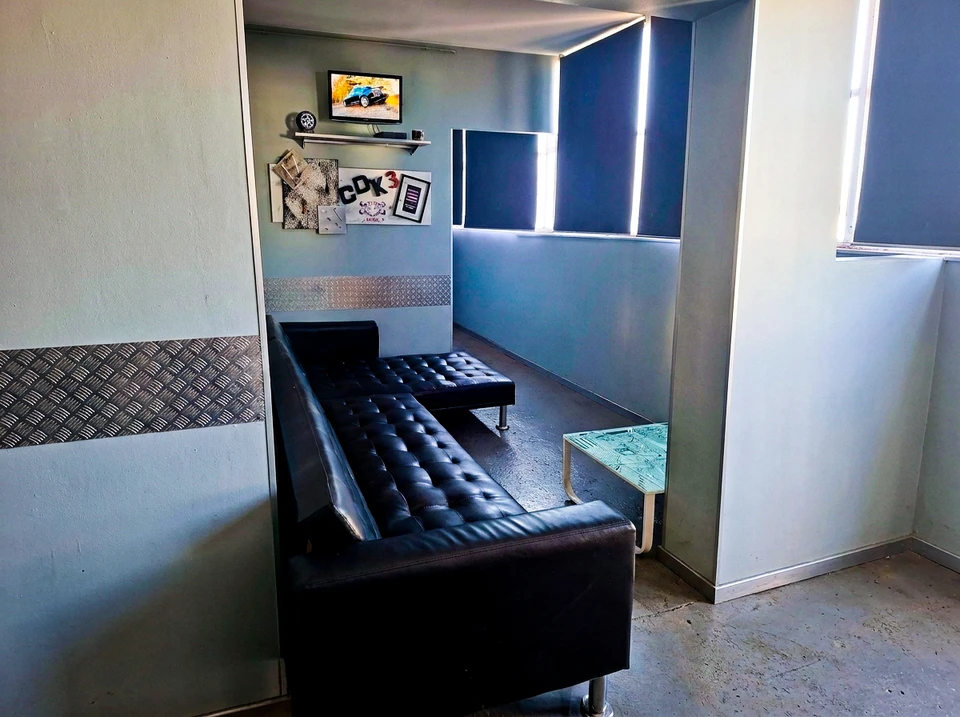 Photographie d'un espace d'attente moderne et accueillant dans un garage automobile. La scène montre un canapé confortable en gris ou bleu clair, une table basse en bois avec une tasse de café fumante servie sur une soucoupe, une lampe douce en arrière-plan, des plantes vertes discrètes, et à travers une vitre, une vue partielle de l'atelier de mécanique avec les véhicules en cours de révision. L'atmosphère est chaleureuse, lumineuse et professionnelle, incarnant l'accueil bienveillant et le confort client.