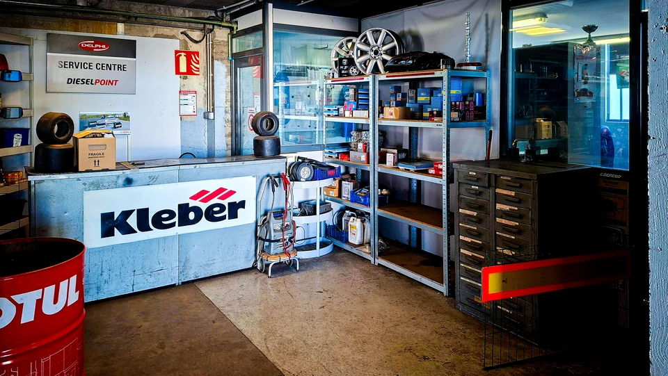 Photo professionnelle d'un espace d'accueil moderne et accueillant dans un garage automobile. L'image montre un canapé ou des sièges confortables en attente, une table basse avec du café ou des boissons, une connexion wifi disponible, et un écran mural affichant les travaux en cours. L'espace est bien éclairé, propre et professionnel, avec un accès visible adapté aux personnes à mobilité réduite. L'atmosphère doit transmettre chaleur, professionnalisme et bien-être client.