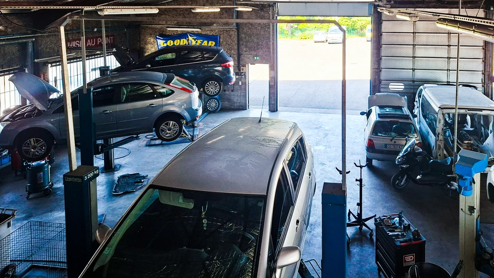 Photographie d'un technicien automobile en combinaison professionnelle effectuant un diagnostic sur une voiture dans un atelier de garage, utilisant un équipement diagnostic professionnel connecté au véhicule, avec des outils de réparation visibles en arrière-plan. La scène capture un moment de travail professionnel concentré, montrant l'expertise et la transparence du diagnostic.