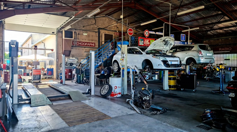 Garage mécanique automobile intérieur, deux véhicules blancs sur ponts élévateurs à colonnes, capot ouvert, moteur déposé au sol sur support